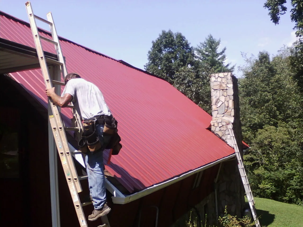 Roofer on a ladder beside a red metal roof and stone chimney — Soffit & Fascia Repair by Roofing Brothers in Lake Saint Louis